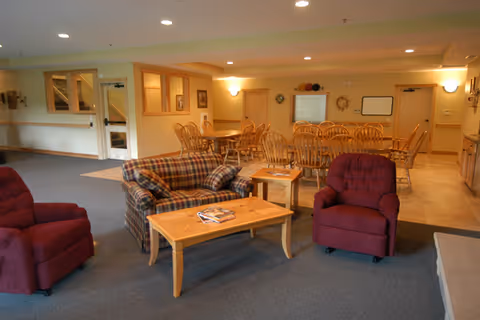 A spacious common area in an assisted living facility featuring a plaid sofa and two red armchairs arranged around a wooden coffee table with magazines. In the background, there are multiple wooden dining tables and chairs, beige walls, and soft overhead lighting.