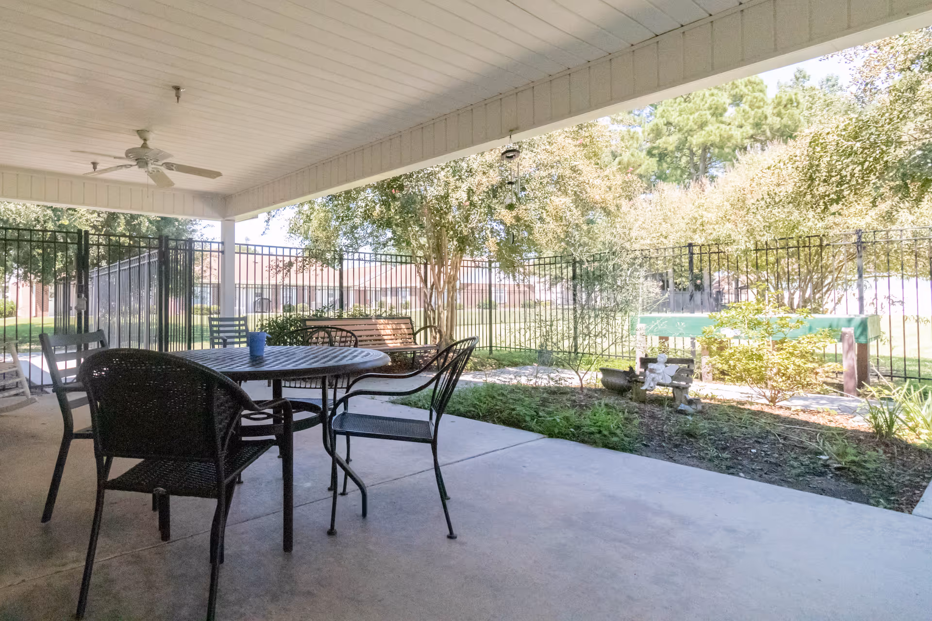 Covered outdoor patio area with a round metal table and several chairs. There is a ceiling fan above and a fenced garden area with trees and shrubs beyond the patio.