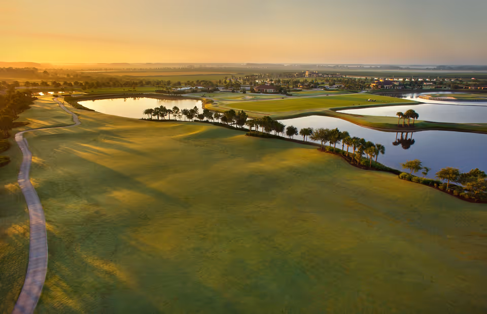 Aerial view of a golf course at sunset with green fairways, water hazards, palm trees, and a winding cart path. The landscape extends to the horizon with buildings and open fields in the distance.