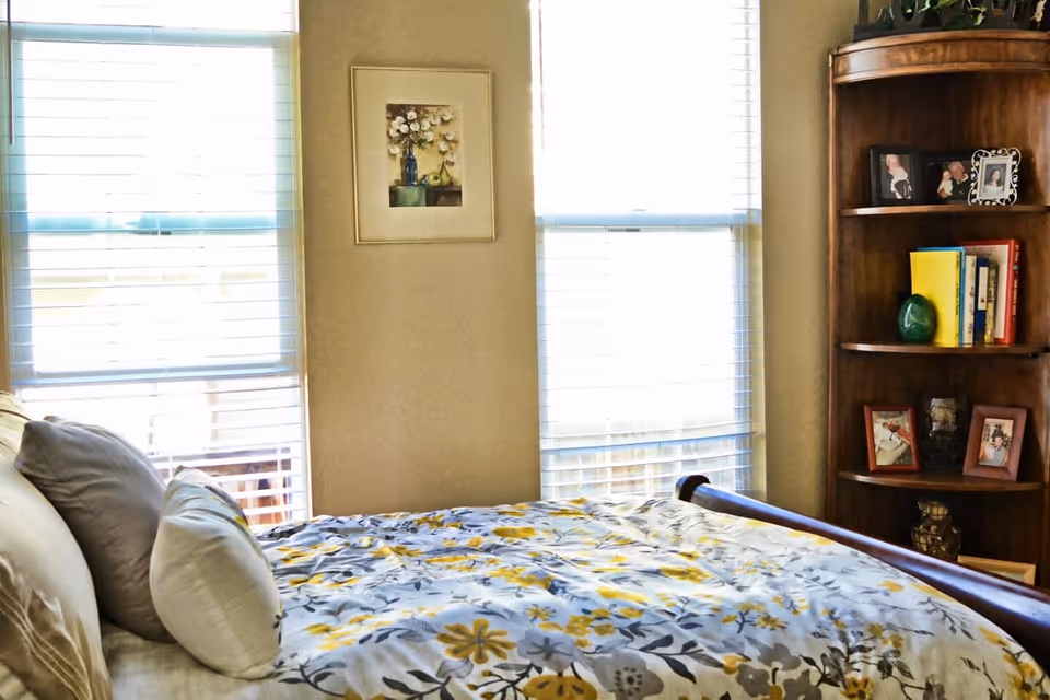 A bedroom with a bed covered in a floral patterned comforter in shades of yellow, gray, and white. The bed has several pillows. Behind the bed are two large windows with white blinds, and a framed floral painting hangs on the wall between the windows. To the right is a wooden corner bookshelf with framed photos, books, and decorative items.