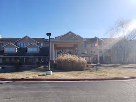 Front exterior of a multi-story senior living building with a covered entrance, flagpole, lamp post, and landscaped lawn under a clear sky.