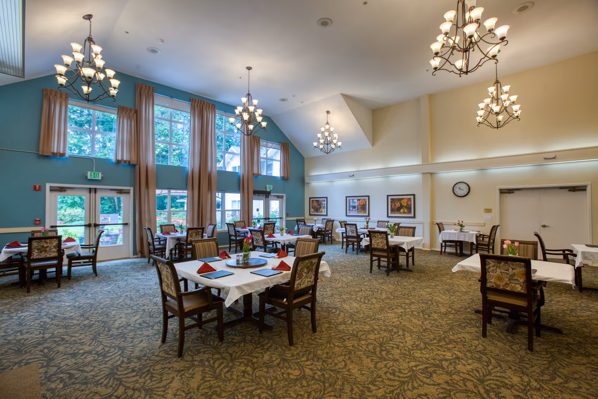 A spacious dining room with multiple tables covered with white tablecloths and set with red napkins. The room features large windows with beige curtains, patterned carpet, several chandeliers hanging from a high ceiling, and framed artwork on the walls. There are chairs around each table and a clock on the far wall.
