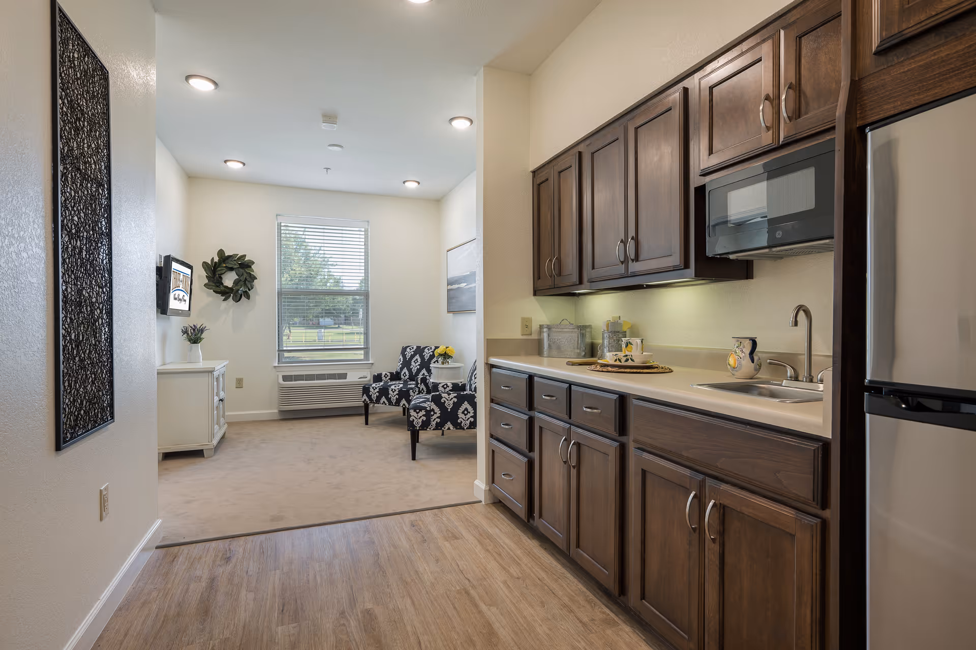 Apartment interior showing a kitchenette with dark wood cabinets and sink, and a small sitting area with patterned chairs by a window.