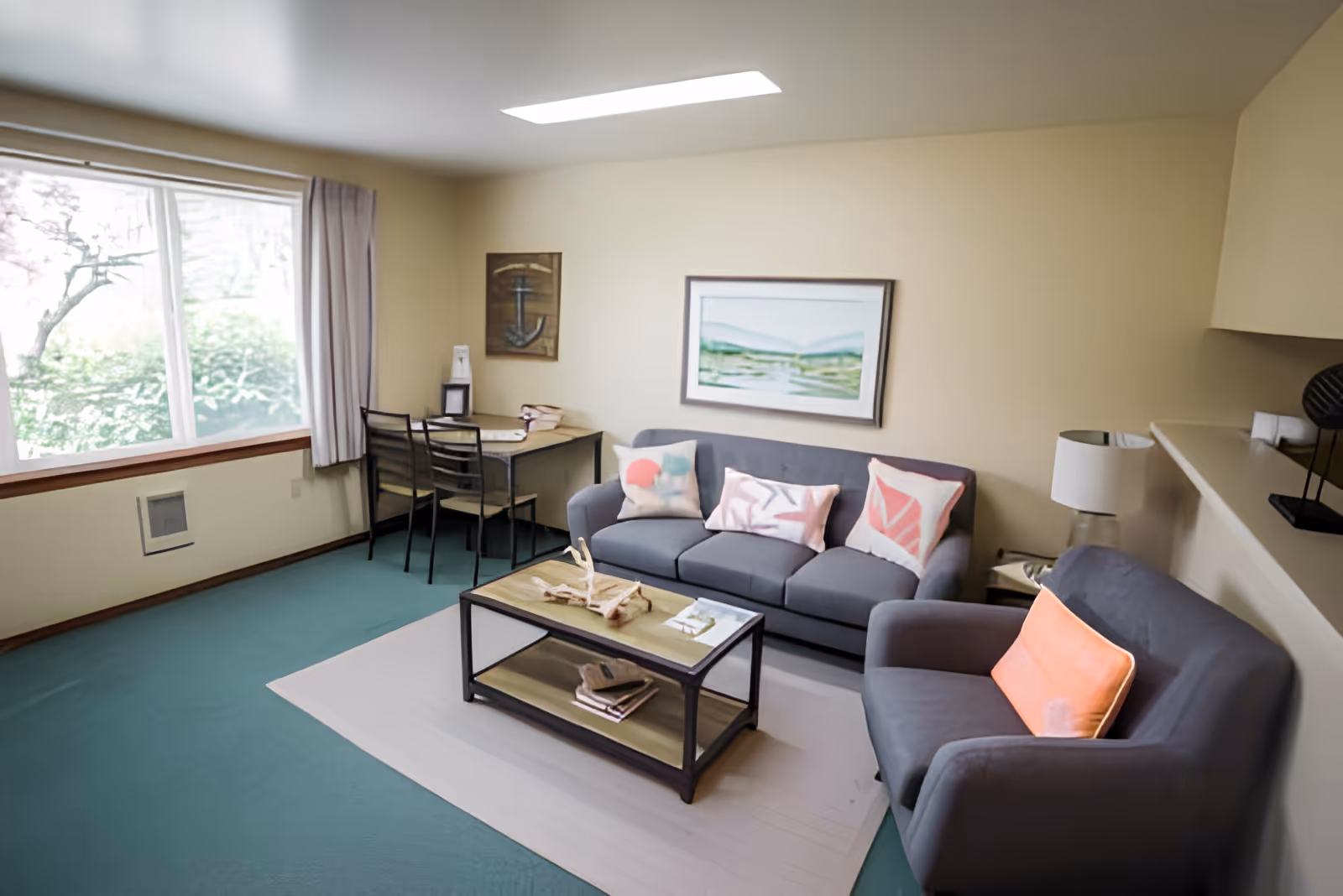 A cozy living room with a gray sofa adorned with three decorative pillows and a matching armchair with an orange pillow. A wooden coffee table with books and decorative items sits on a light-colored rug. A small dining table with two chairs is placed near a large window with curtains, allowing natural light to brighten the room. The walls are beige, decorated with framed artwork, and a lamp is positioned on a side table next to the armchair.