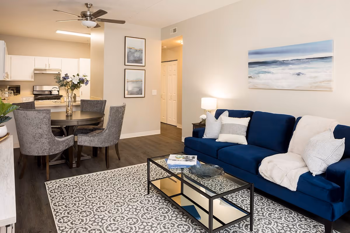 A modern living room and dining area featuring a blue sofa with white and gray pillows and a white throw blanket, a glass coffee table with books and a decorative bowl, a patterned area rug, a round dining table with four gray upholstered chairs, a vase with flowers on the table, white kitchen cabinets, stainless steel appliances, two framed abstract paintings on the wall, a ceiling fan, and a hallway leading to other rooms.