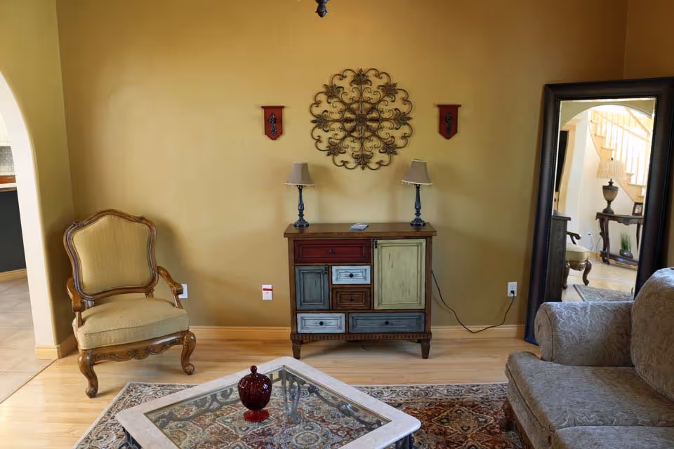 A cozy living room area with a vintage armchair on the left, a multicolored wooden cabinet with two table lamps on top against a yellow wall, and a large ornate mirror on the right reflecting a staircase and part of another room. A patterned rug and a glass-top coffee table with a decorative red item are in the foreground.