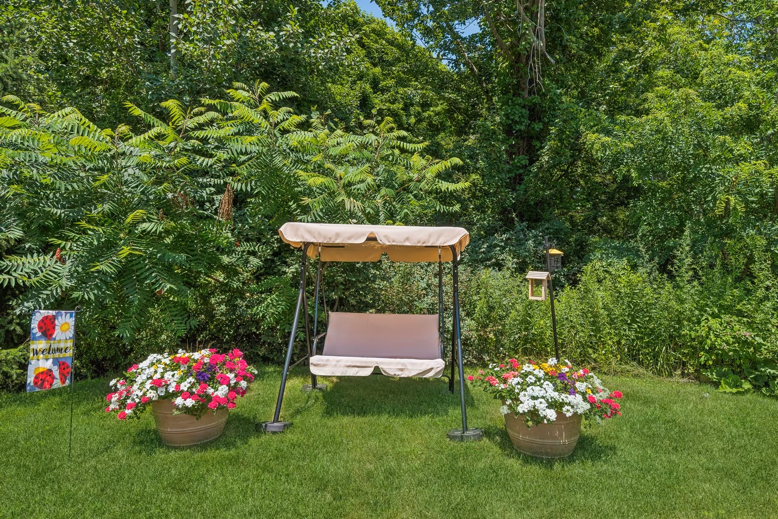 A canopy swing bench centered on a lawn between two large flower pots with colorful blooms, a bird feeder, and dense green trees in the background.