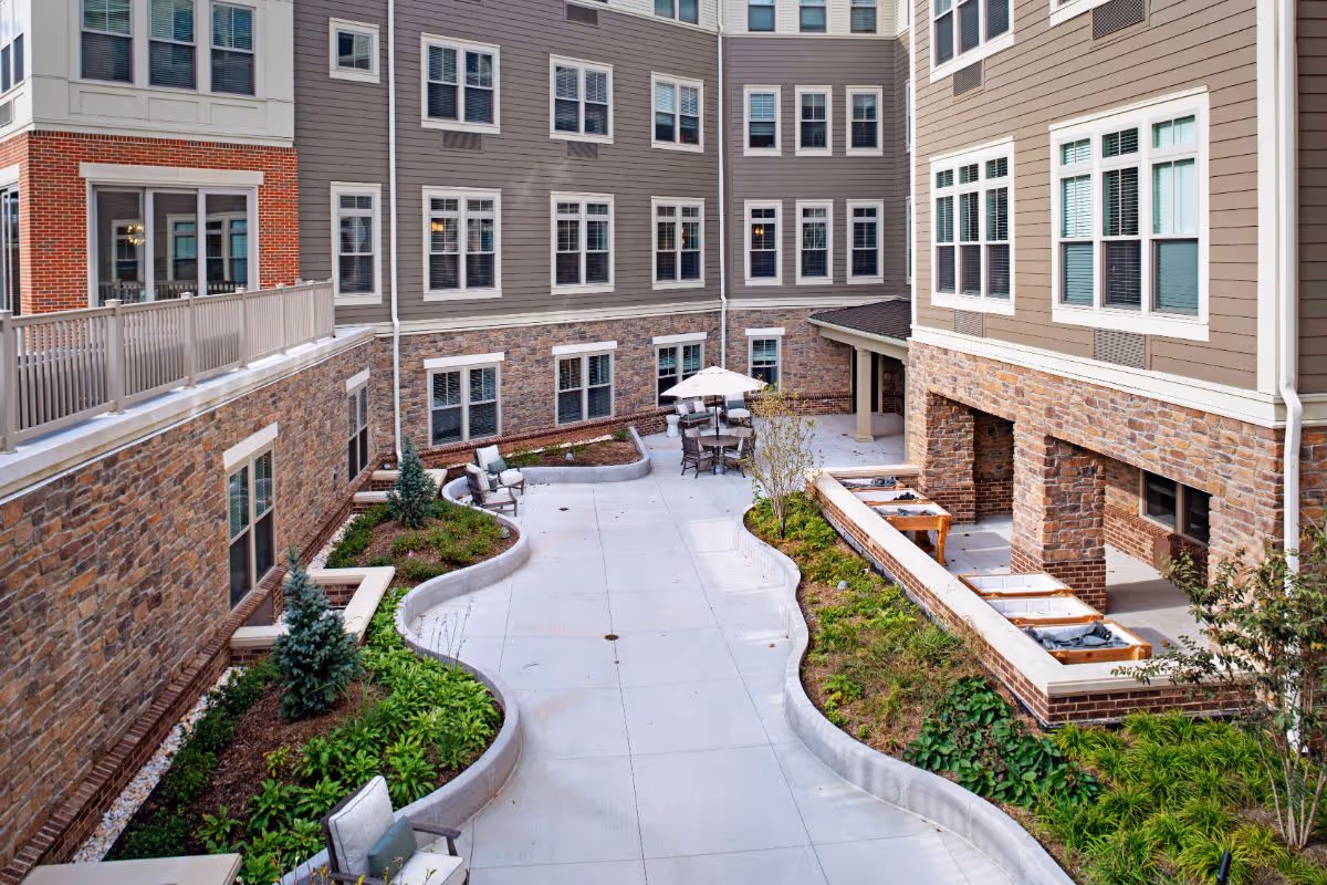 Outdoor courtyard area of a senior living facility with curved garden beds, small trees, and shrubs. There are several chairs and tables with umbrellas arranged on a concrete patio, surrounded by multi-story buildings with stone and siding exteriors and many windows.