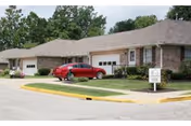 Exterior view of a single-story brick residential building with attached garages and a red car parked in front. The building is surrounded by a well-maintained lawn and trees in the background. A sign is visible near the sidewalk indicating the name of the facility.
