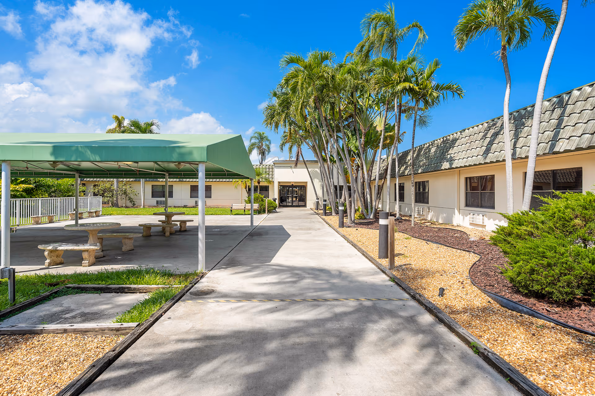 Outdoor view of Shoreside Health and Rehabilitation Center showing a paved walkway leading to the entrance of a single-story building with a tiled roof. On the left side, there is a covered seating area with stone benches and tables. Palm trees and landscaped bushes line the right side of the walkway under a bright blue sky with some clouds.