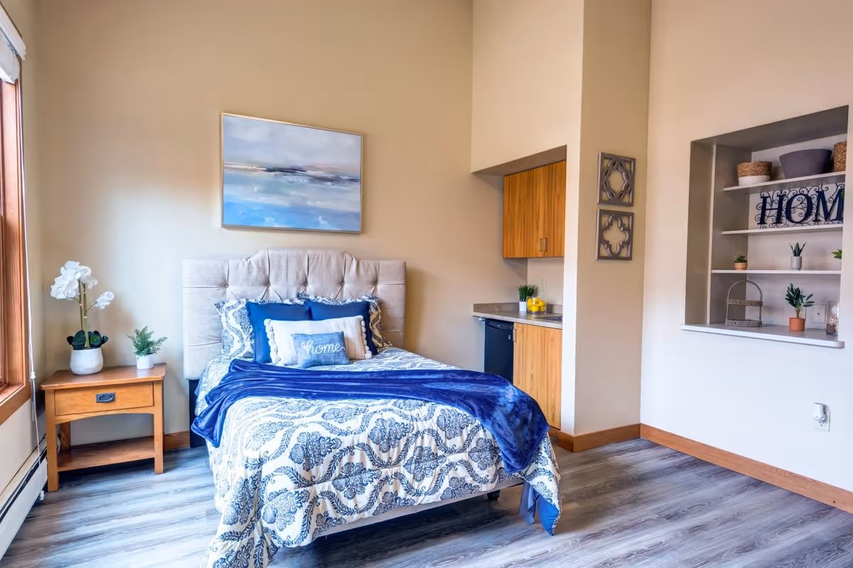 A cozy bedroom with a bed featuring a tufted beige headboard, blue and white patterned bedding, and decorative pillows including one with the word 'home'. To the left of the bed is a wooden nightstand with two potted plants. Above the bed hangs a landscape painting with blue and gray tones. On the right side, there is a small kitchenette area with wooden cabinets and a countertop, and a built-in wall shelf decorated with plants, baskets, and a sign that says 'HOME'. The room has light-colored walls and wood-look flooring.