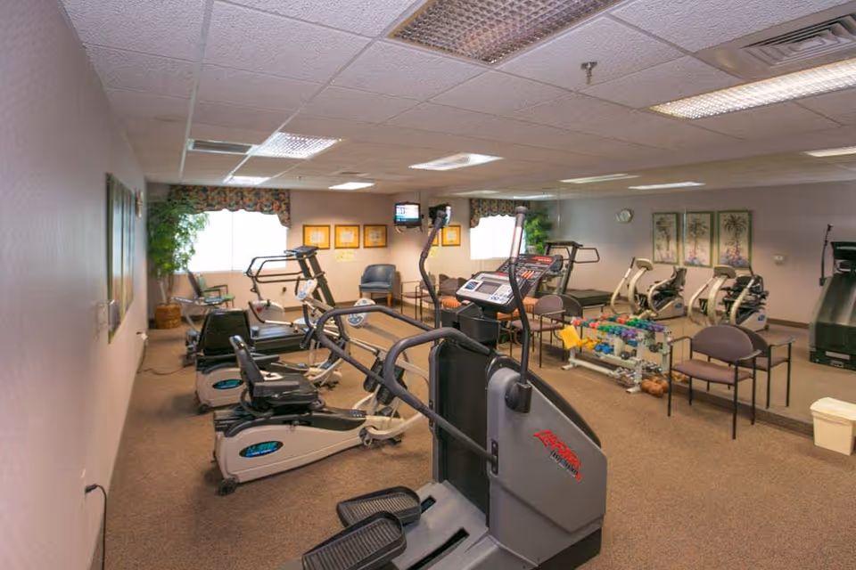 Indoor fitness room with various exercise equipment including treadmills, stationary bikes, and weight racks. The room has beige carpet, mirrored wall, fluorescent ceiling lights, and framed artwork on the walls. There are also chairs and a small TV mounted on the wall.