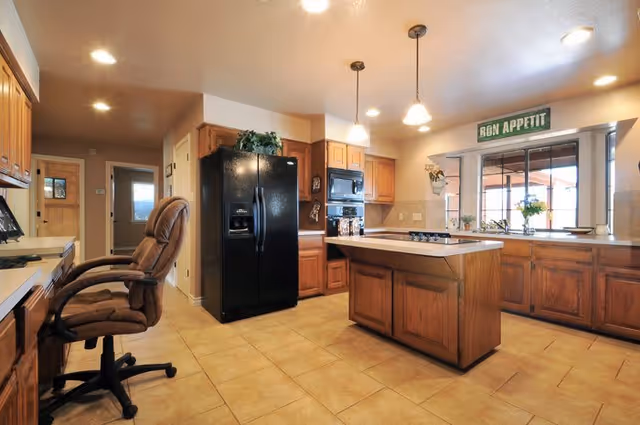 A spacious kitchen with wooden cabinets and a tiled floor. The kitchen features a black refrigerator, built-in microwave, and oven. There is a central island with a cooktop and two pendant lights hanging above it. A large window with a 'Bon Appetit' sign above it lets in natural light. A brown office chair is positioned near a counter on the left side.