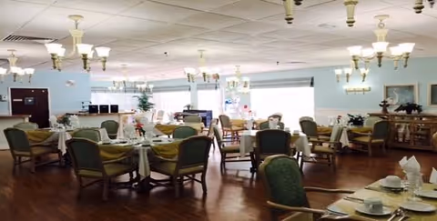 Dining room with multiple round tables set for meals, green upholstered chairs, chandeliers, and large windows.