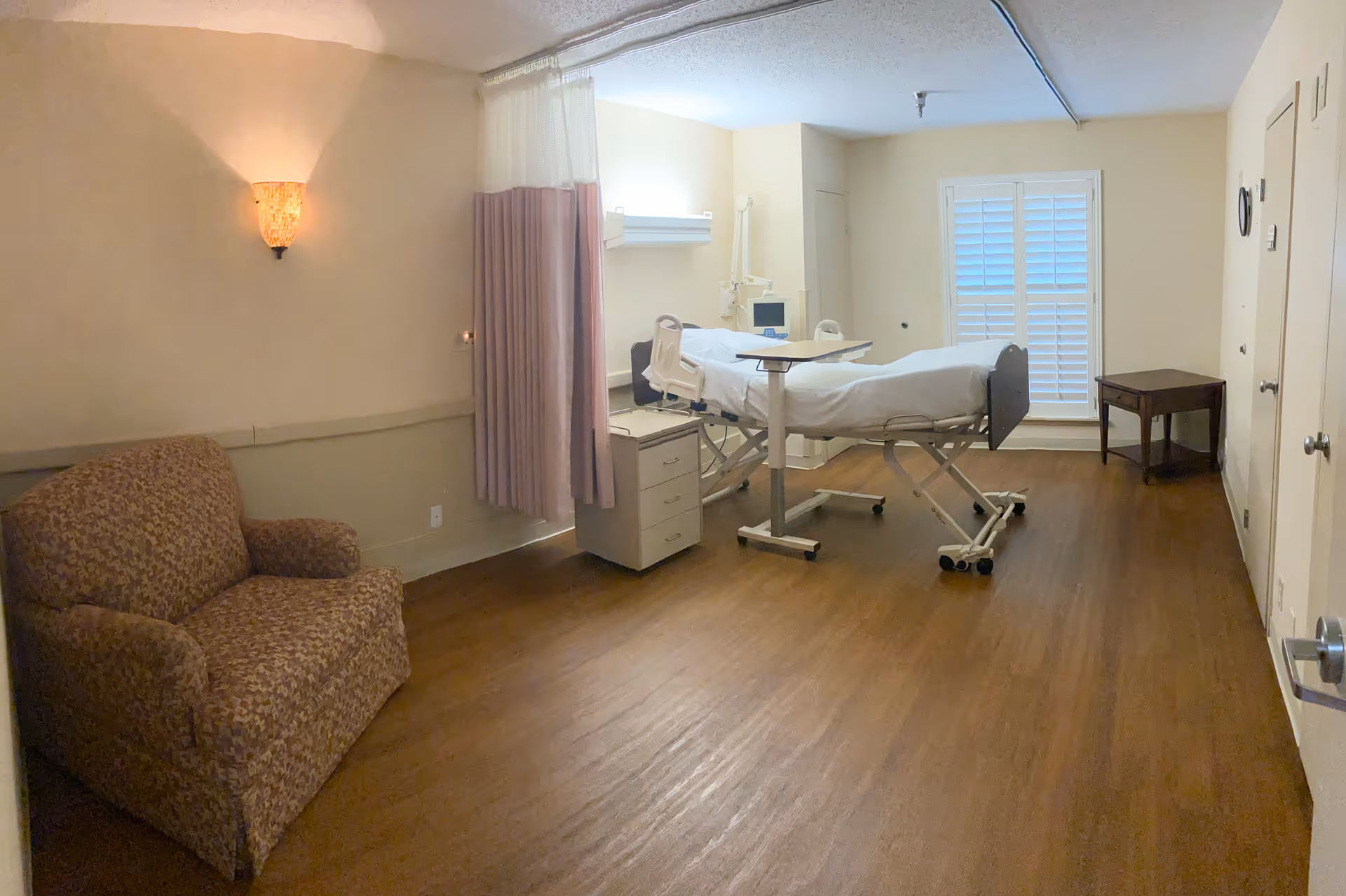 A private patient room with an adjustable hospital bed, privacy curtain, armchair and side tables on wood flooring.