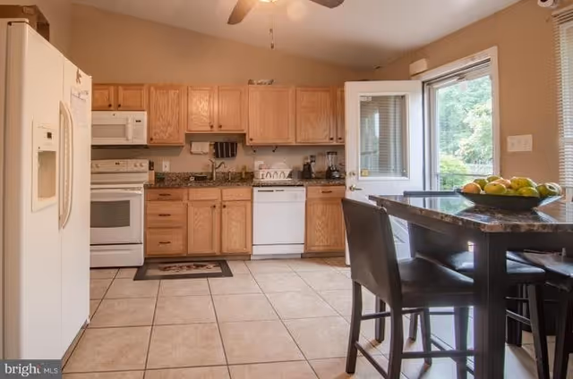 A kitchen with light wood cabinets, a white refrigerator, stove, and dishwasher. There is a granite countertop with a sink and various kitchen appliances. A small dining table with four dark chairs is positioned near a door that opens to an outdoor area with greenery visible through the window.
