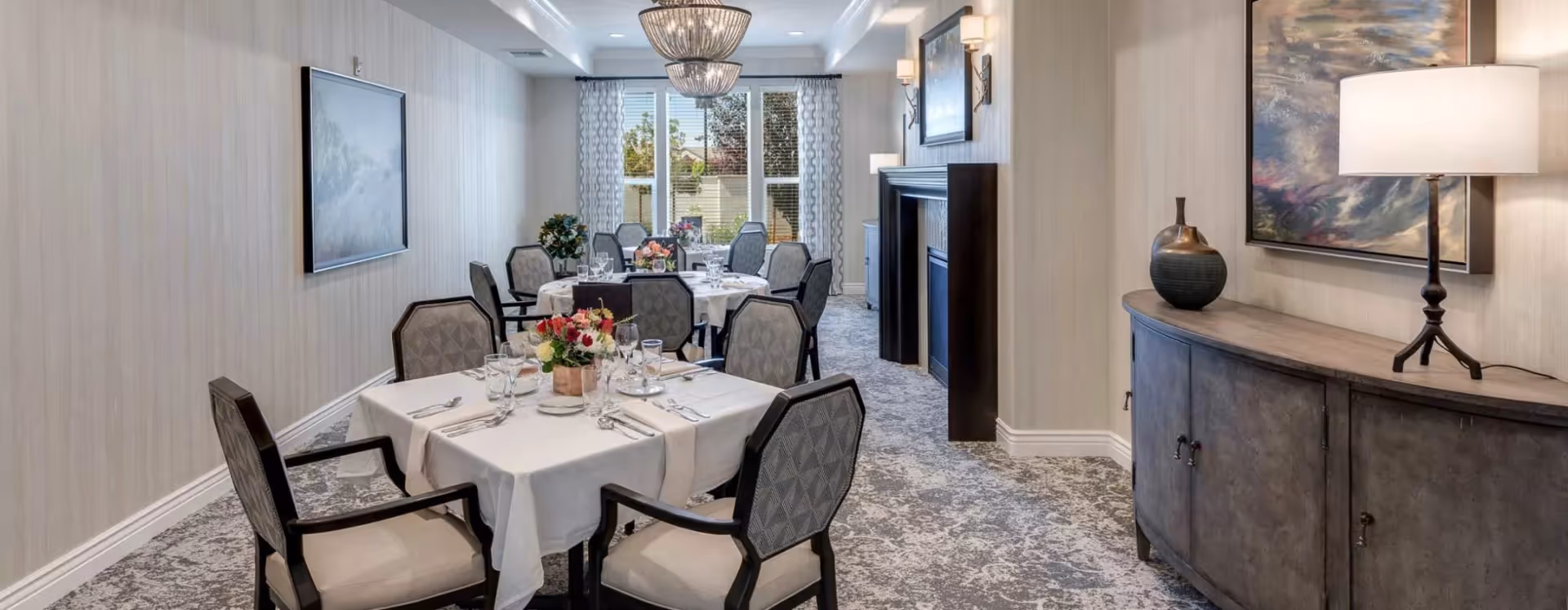A bright dining room with several tables set for a meal, upholstered chairs, chandeliers, and a sideboard.