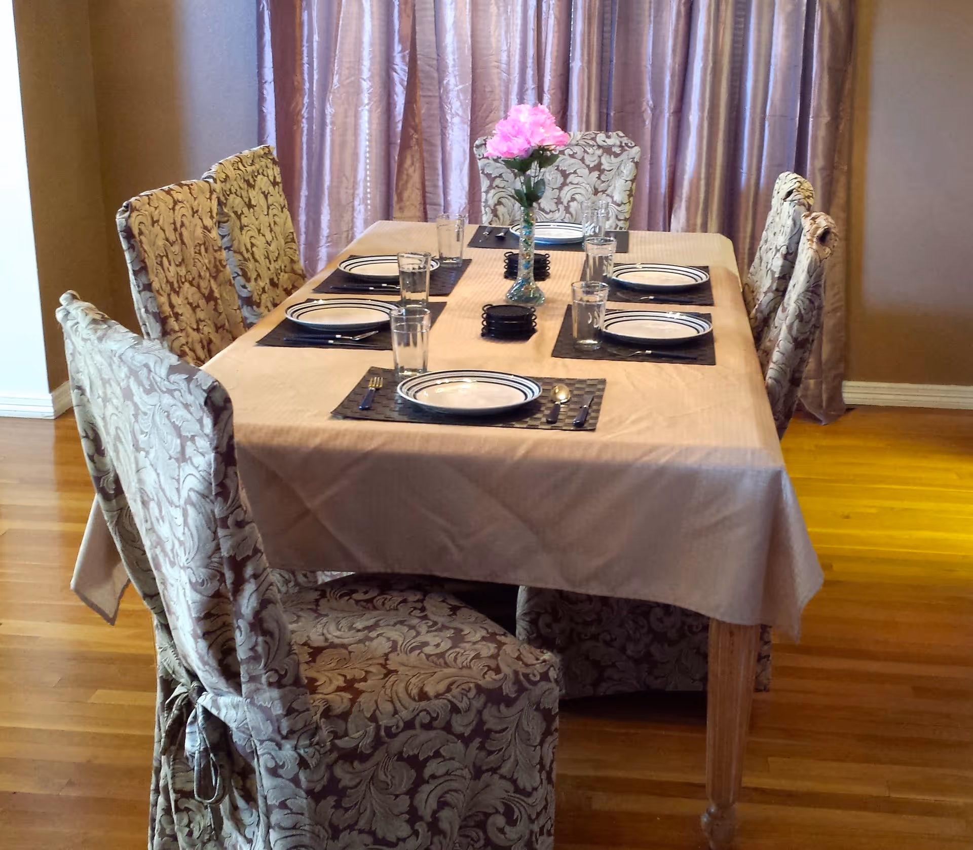 A dining table set for six with patterned slipcovered chairs, place settings and a vase of pink flowers in a room with wood floors and curtains.