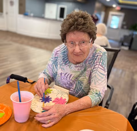 An elderly woman with glasses and a patterned shirt is sitting at a table painting colorful flowers on a paper plate. There is a pink cup with a paintbrush in it and an orange plate on the table. The background shows a spacious room with another person sitting and some furniture.