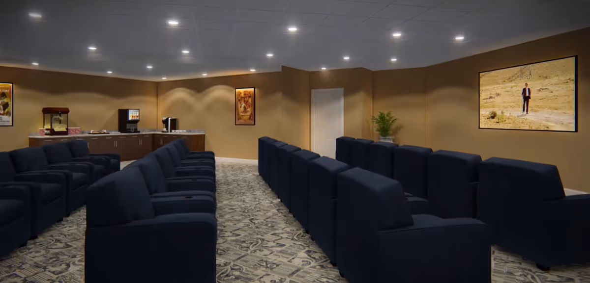 A theater room with multiple rows of dark blue recliner chairs facing a large screen on the wall. The room has beige walls, a patterned carpet, recessed ceiling lights, and a refreshment area with a popcorn machine and beverage dispenser in the back corner.