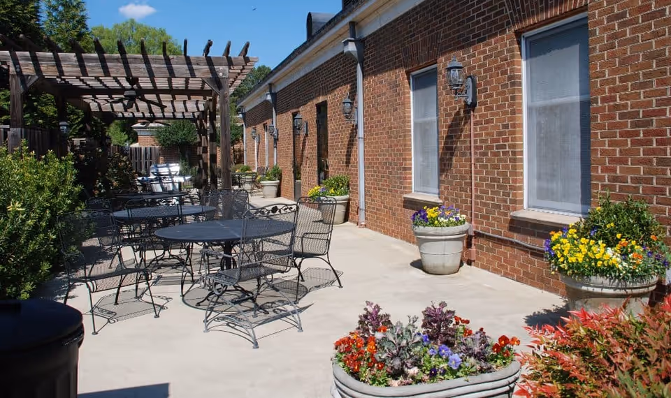 Outdoor patio area with metal tables and chairs, surrounded by potted flowers and plants. A wooden pergola provides partial shade over some seating. The patio is adjacent to a brick building with windows and wall-mounted lantern lights.