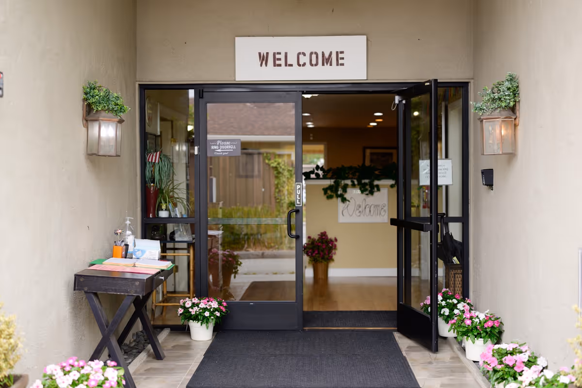 Open glass doors lead into a senior living facility entrance with a Welcome sign and potted plants outside.