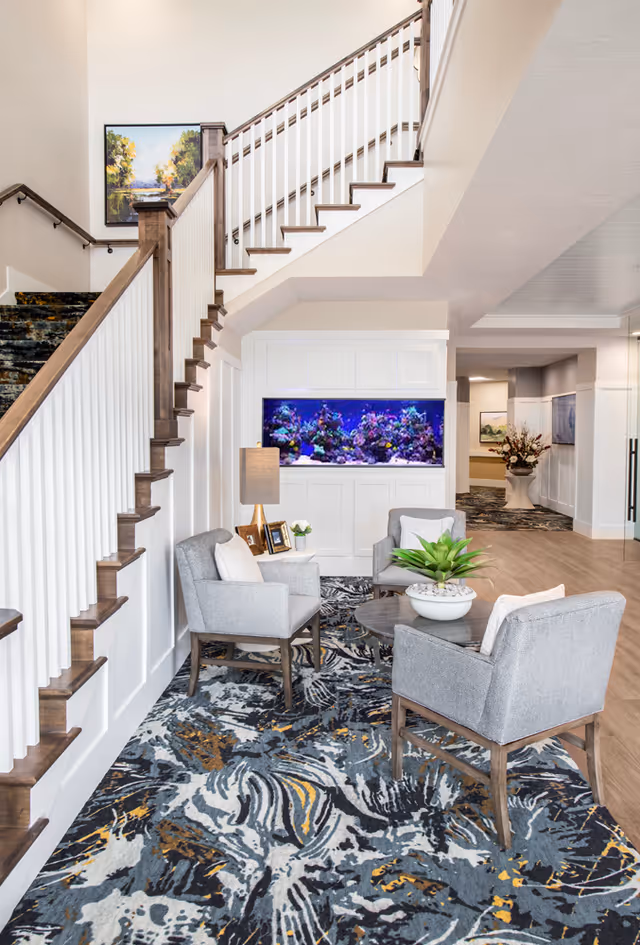 A cozy seating area in a senior living facility featuring three gray upholstered chairs around a round wooden table with a potted plant. Behind the seating area is a built-in aquarium with colorful coral and fish. A staircase with wooden handrails and white balusters ascends on the left side. The floor is covered with a patterned carpet in shades of gray, blue, and gold. A table lamp and framed photos are on a small side table next to one of the chairs. The walls are painted white with wainscoting, and a framed landscape painting hangs above the staircase.
