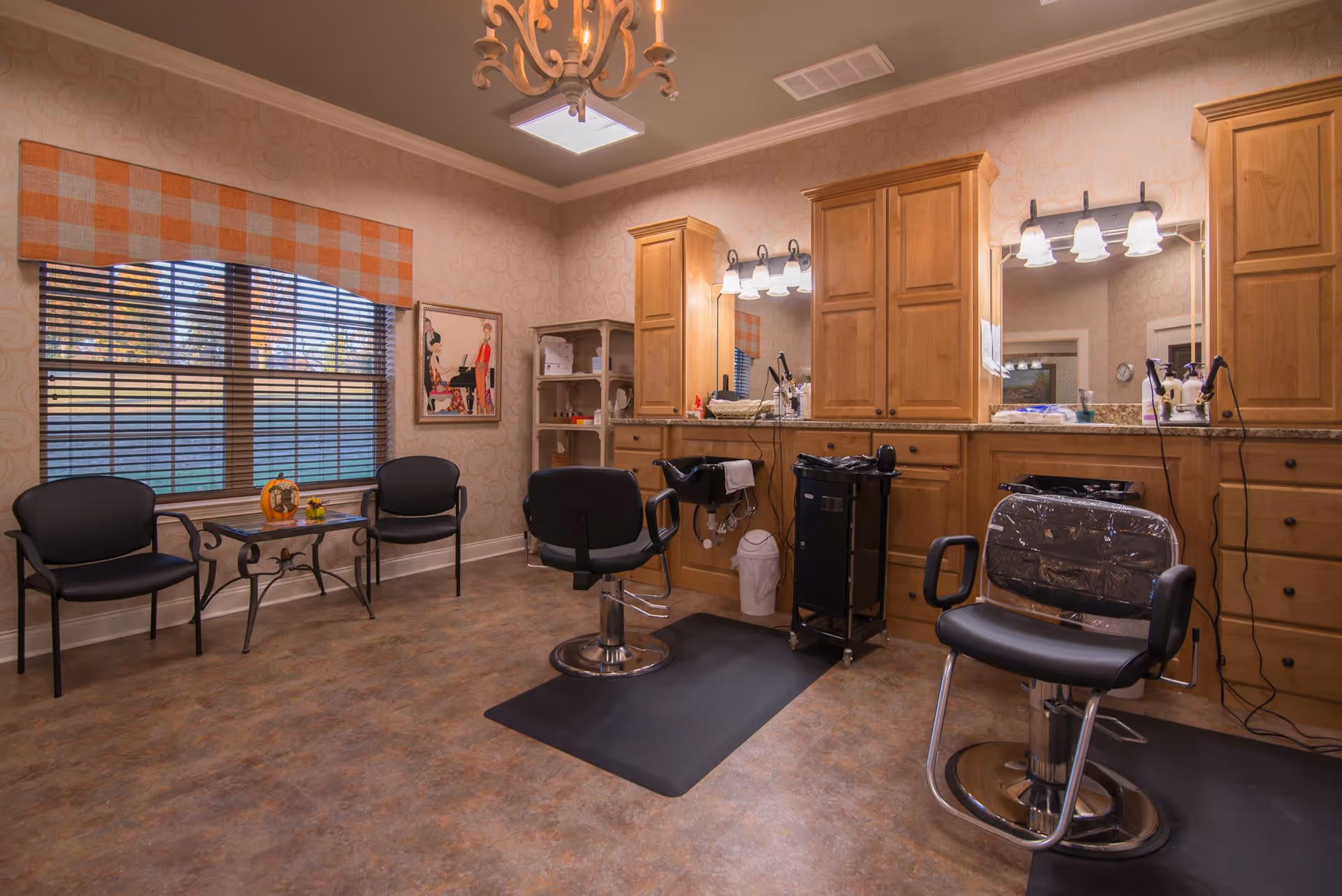 Interior view of a hair salon area in a senior living facility with two black salon chairs in front of large mirrors and wooden cabinets. There are two black waiting chairs and a small table with decorative items near a large window with orange and white checkered valance. The room has warm lighting and patterned wallpaper.