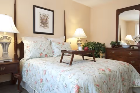 A cozy bedroom with a floral-patterned bedspread and matching pillows on a wooden bed frame. There is a wooden tray on the bed, two bedside tables each with a lamp, a framed picture on the wall above the bed, a wooden dresser with a large mirror, and a green potted plant beside the dresser.