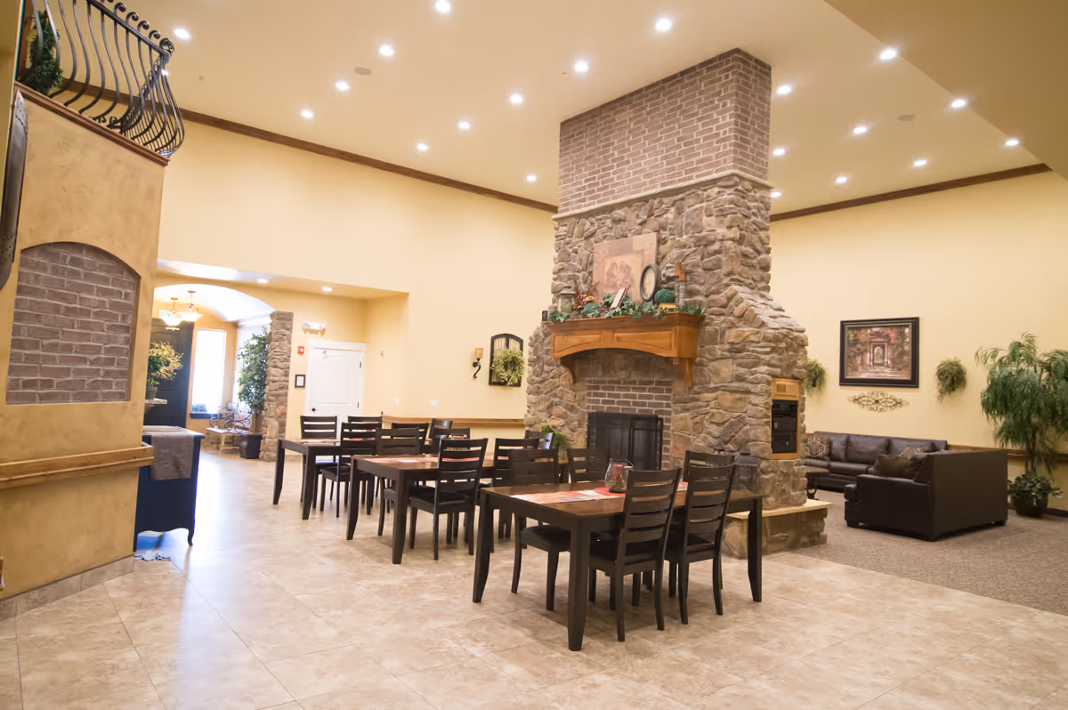 A spacious interior common area featuring a large stone fireplace in the center, surrounded by wooden tables and chairs. To the right, there is a seating area with dark brown sofas and green plants against a beige wall with framed artwork. The floor is tiled, and the ceiling is fitted with recessed lighting.