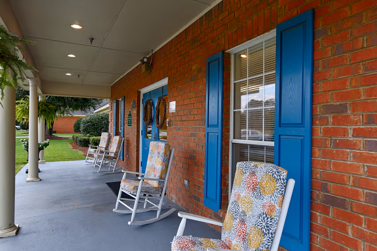 Covered porch area of a brick building with blue window shutters and blue double doors. Several cushioned rocking chairs with floral patterns are arranged along the porch. There are white columns supporting the porch roof and green plants hanging and placed in pots. A well-maintained lawn and bushes are visible in the background.