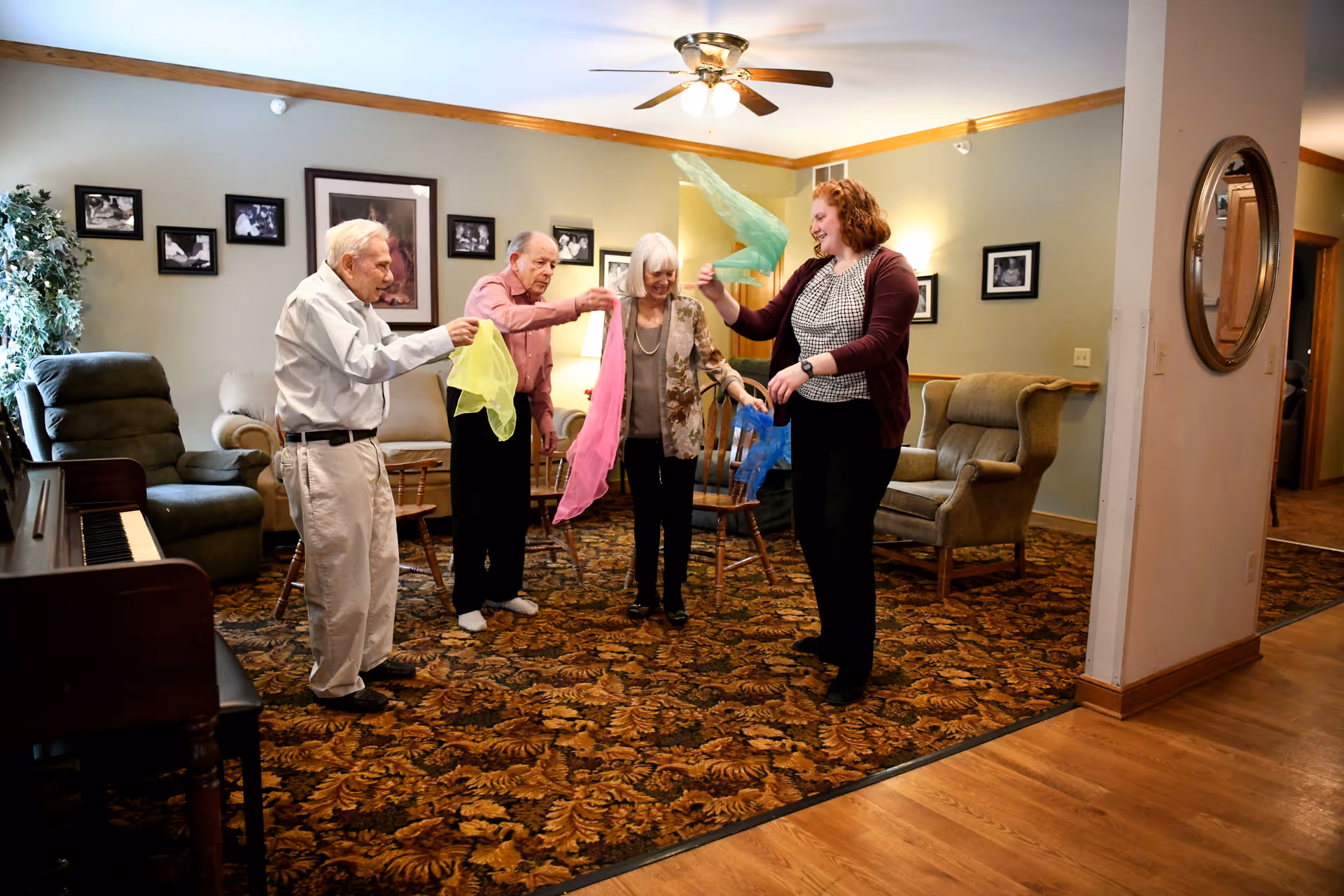 A group of three elderly people and a younger woman standing in a living room area, playing with colorful scarves. The room has patterned carpet, comfortable chairs, framed photos on the wall, a piano, and a ceiling fan with lights.