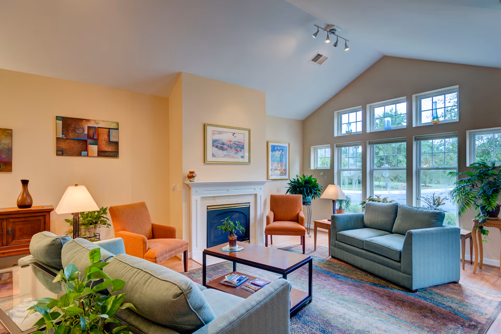 A bright and spacious living room with high vaulted ceilings and large windows letting in natural light. The room features two green sofas, two orange armchairs, a wooden coffee table, a fireplace with framed artwork above it, several potted plants, and decorative lamps. The walls are painted a soft beige color.