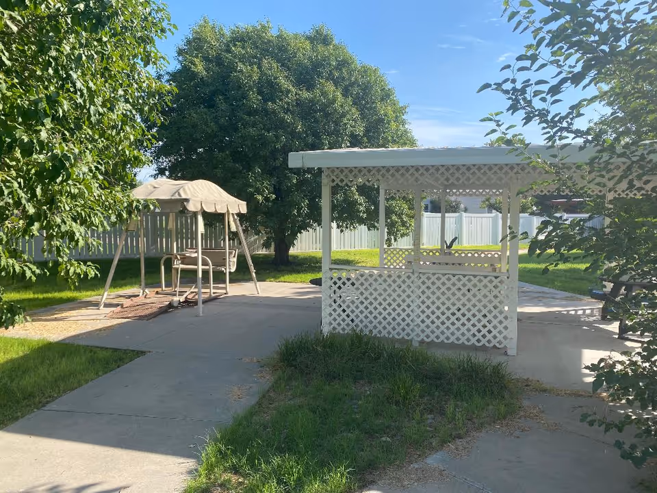 Outdoor area with a white lattice gazebo and a covered swing set on a concrete patio surrounded by green grass and trees under a clear blue sky.