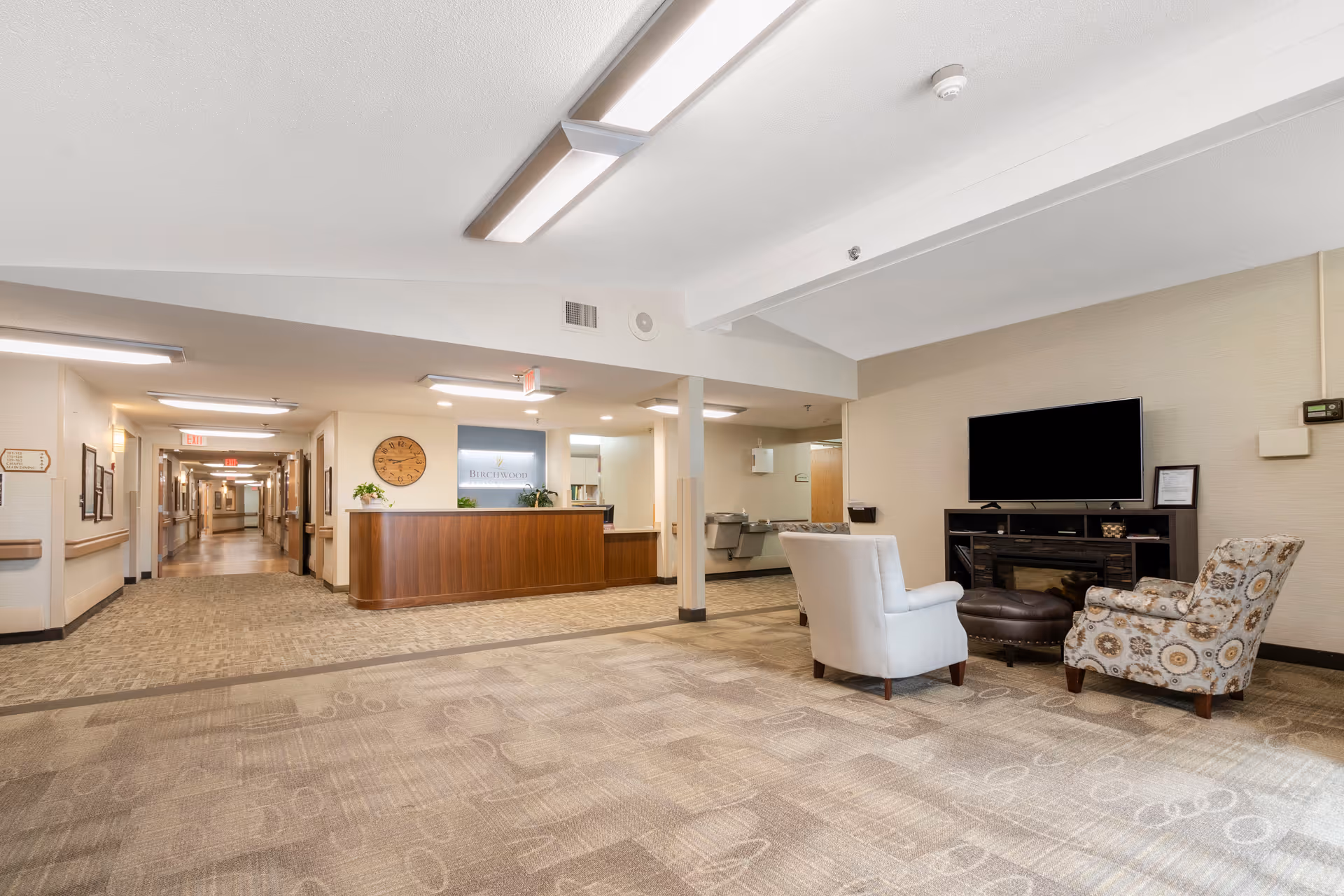 Interior view of Birchwood Healthcare Center SNF showing a reception desk with a clock on the wall behind it, a hallway extending to the left, and a seating area with two armchairs and a large flat-screen TV on a stand to the right.