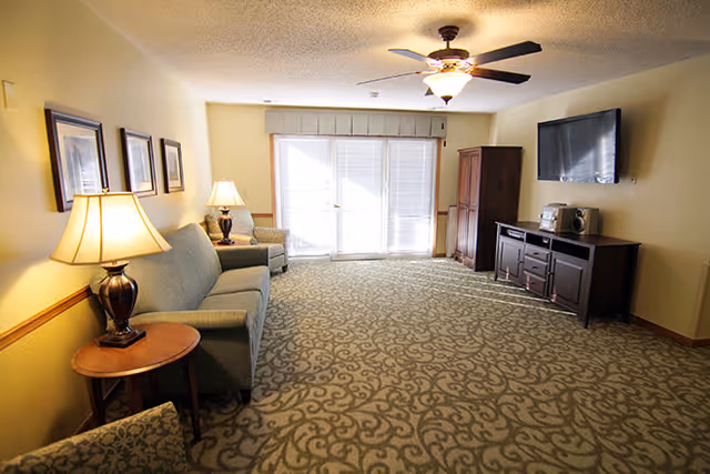 Carpeted living room with sofas and side tables with lamps, a wall-mounted TV, wooden cabinet, and glass doors letting in natural light.