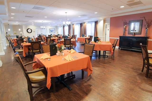Spacious dining room with multiple tables covered in orange tablecloths, set with folded napkins and floral centerpieces, and wooden chairs on hardwood floors.