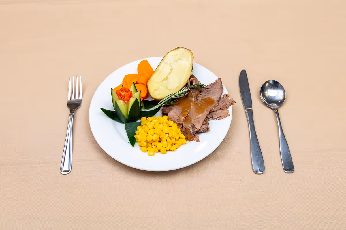 A white plate with a serving of sliced roast beef with gravy, a baked potato half, cooked corn, carrot slices, and a cucumber garnish shaped like a flower on a beige tablecloth. A fork is placed to the left of the plate, and a knife and spoon are placed to the right.