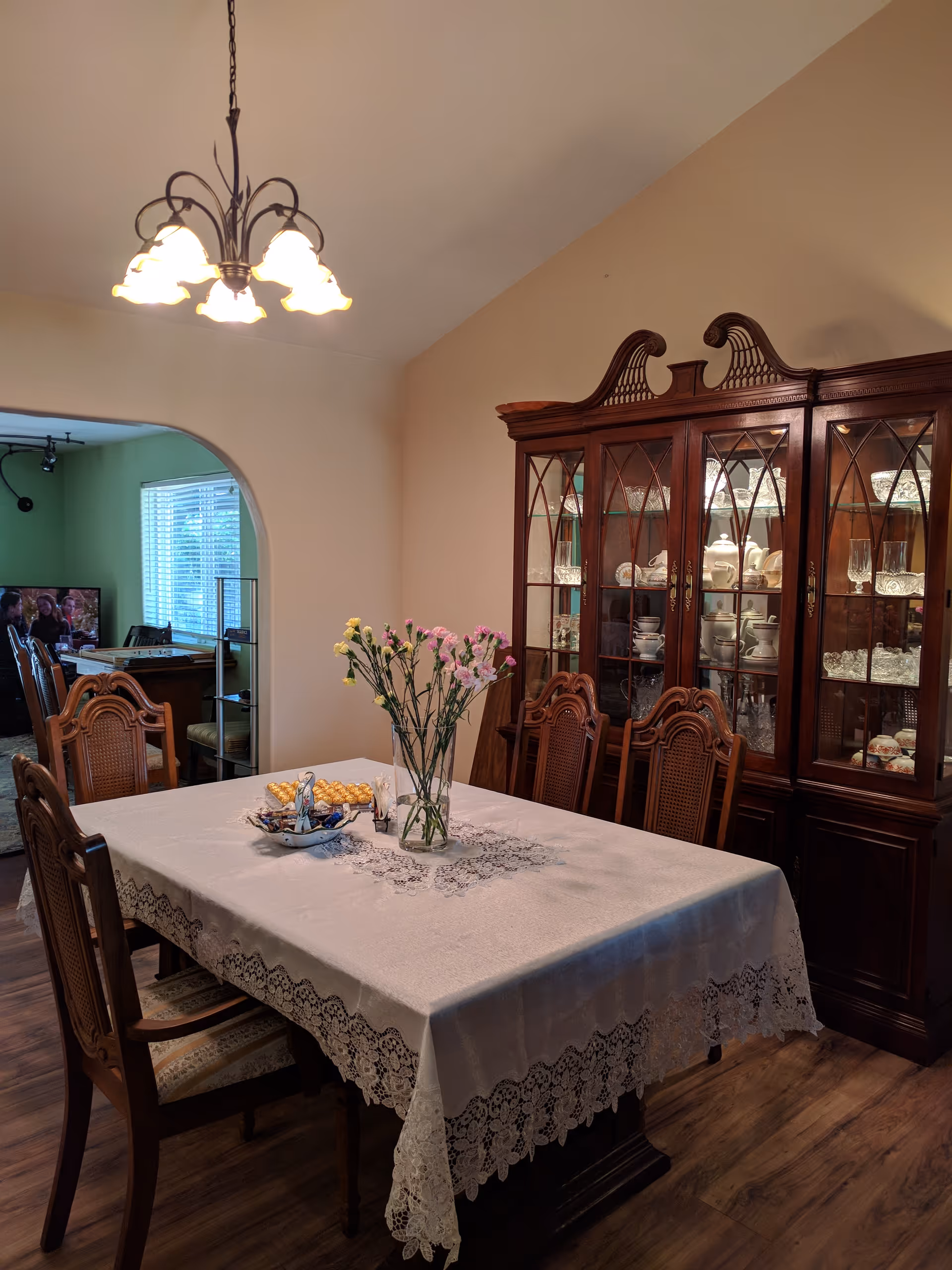 A dining room with a wooden dining table covered with a white lace tablecloth. There are six wooden chairs around the table. A glass vase with pink and yellow flowers is placed in the center of the table along with a decorative dish. Behind the table is a large wooden china cabinet filled with glassware and dishes. A chandelier with multiple lights hangs from the ceiling. An archway leads to another room where a television and additional furniture are visible.