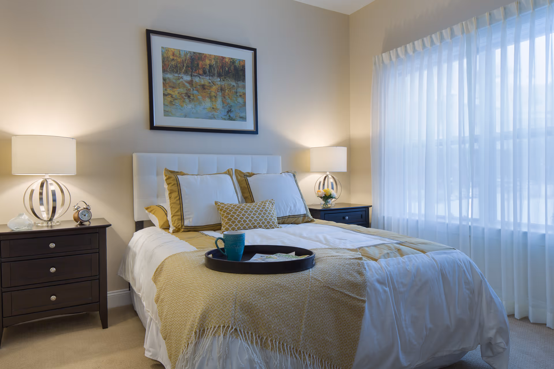A neatly made bed with white and gold bedding in a bright bedroom. There are two bedside tables with modern lamps on each side of the bed. A framed painting hangs above the headboard, and sheer white curtains cover a large window letting in natural light. A tray with a blue mug and some papers is placed on the bed.