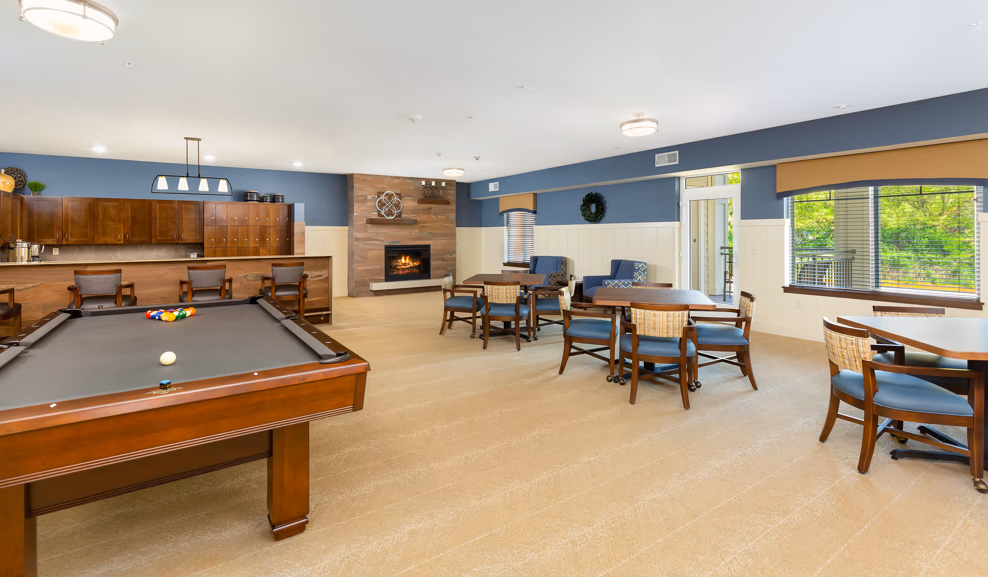 A spacious common area with a pool table in the foreground, several tables and chairs arranged for seating, a fireplace with a wooden accent wall, and a counter with bar stools. Large windows let in natural light and offer a view of greenery outside.
