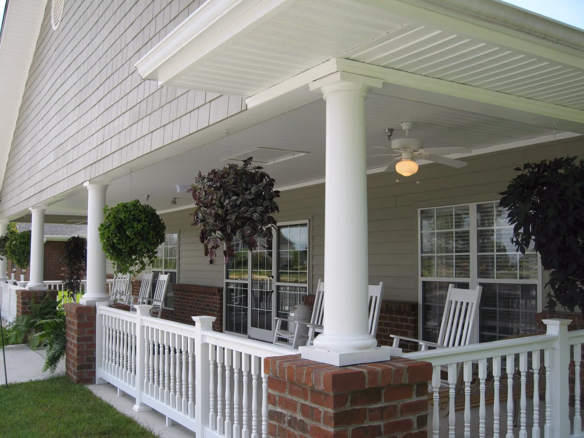 Covered outdoor porch area with white rocking chairs, white railing, brick pillars, hanging plants, ceiling fans, and windows with white frames.