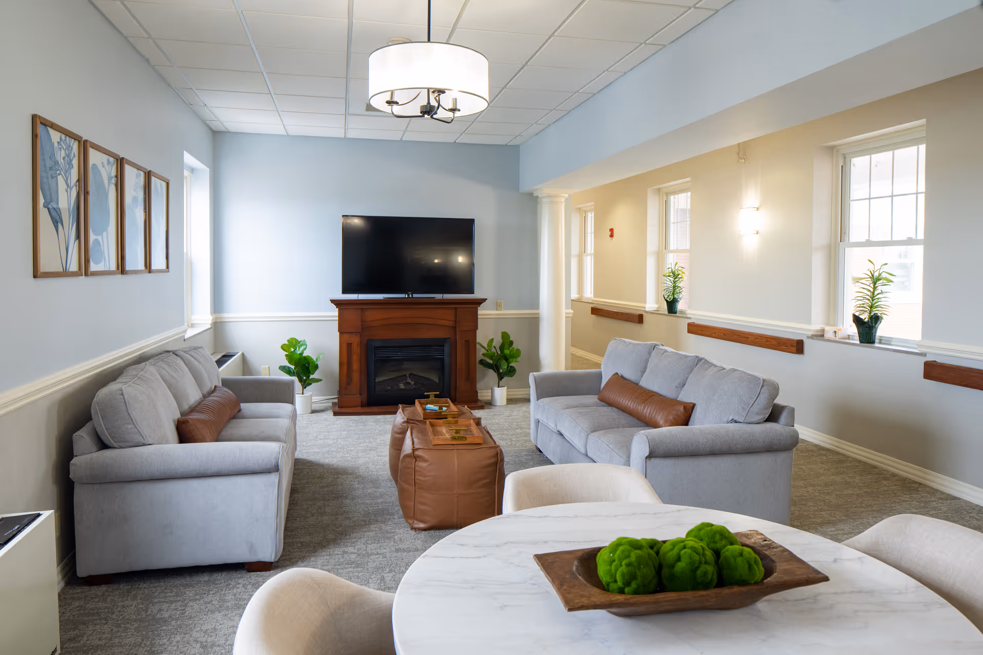 A cozy senior living common area with two gray sofas facing each other, a wooden fireplace with a flat-screen TV mounted above it, a round white marble table with a decorative wooden tray holding green moss balls, and several potted plants near the windows and fireplace. The room has light blue and beige walls, carpeted floor, and a modern ceiling light fixture.