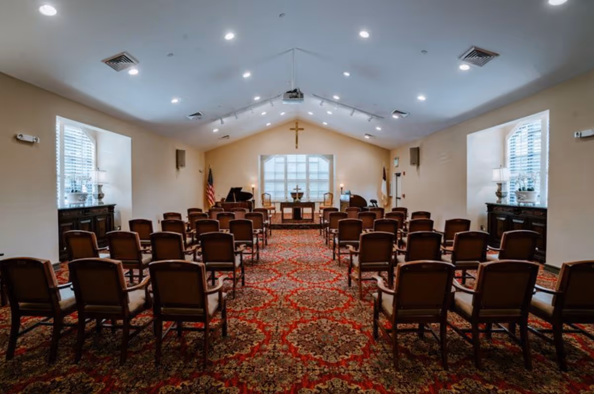 Carpeted chapel-style meeting room with rows of chairs facing a podium and cross on the far wall.
