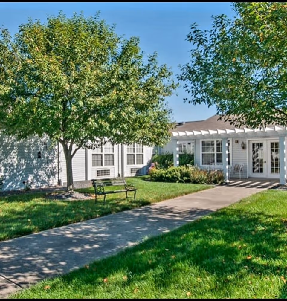 Outdoor view of a senior living facility with a concrete walkway leading to a white building with large windows and a pergola-covered entrance. There are green trees, a bench, and well-maintained grass surrounding the path under a clear blue sky.