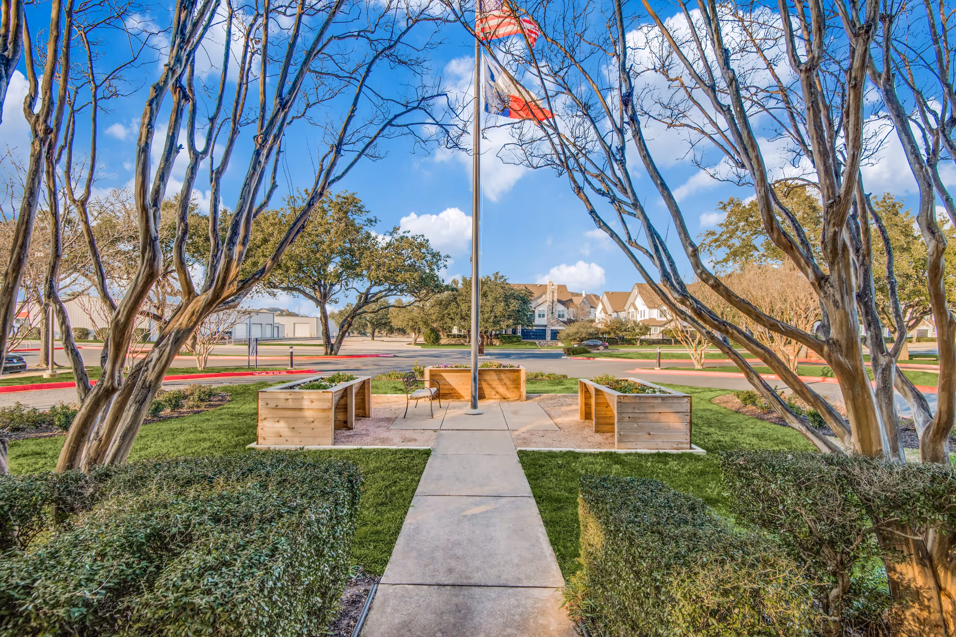 Outdoor garden area with a concrete pathway leading to a flagpole flying the American and Texas flags. The area is surrounded by neatly trimmed bushes and trees with bare branches. There are wooden planter boxes on either side of the flagpole and a single metal chair near the center. In the background, there is a parking lot and residential-style buildings under a blue sky with scattered clouds.
