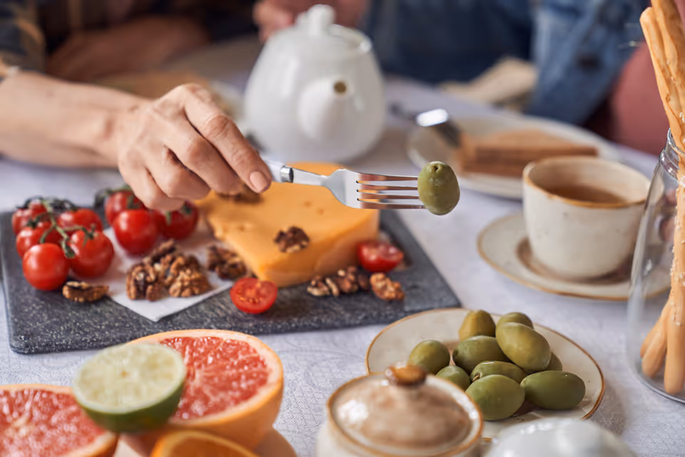 A close-up of a hand holding a fork with a green olive on it over a table set with various foods including cherry tomatoes, walnuts, a block of cheese, halved citrus fruits, a cup of coffee, a teapot, and breadsticks in a jar.