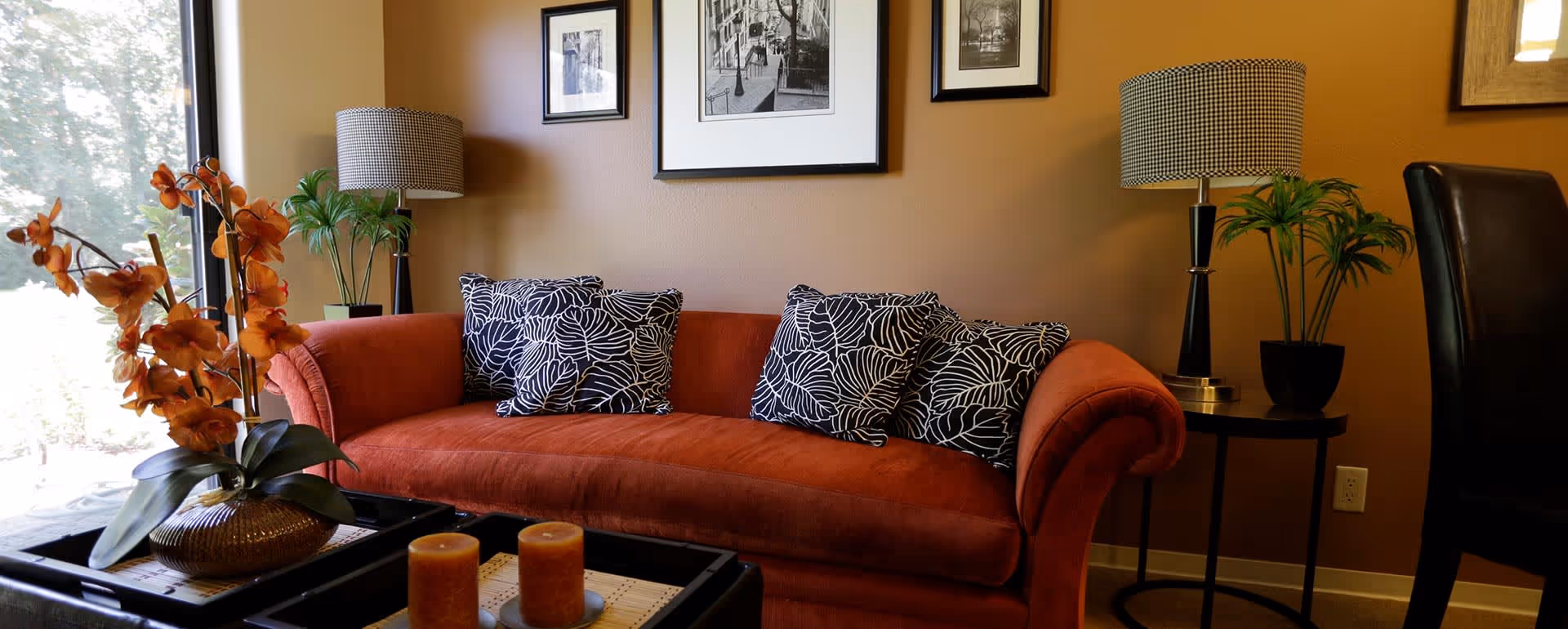 A cozy living room area with a burnt orange sofa adorned with three black and white patterned pillows. There is a black coffee table in front of the sofa with two orange candles and a decorative plant with orange flowers. On either side of the sofa are small round tables with black bases, each holding a lamp with a patterned shade and a green potted plant. The wall behind the sofa is painted a warm brown and decorated with three framed black and white photographs. A large window on the left side lets in natural light.