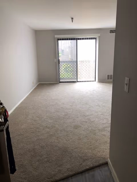 Empty room with beige carpet flooring and white walls, featuring a sliding glass door with vertical blinds leading to a small balcony with a lattice railing.