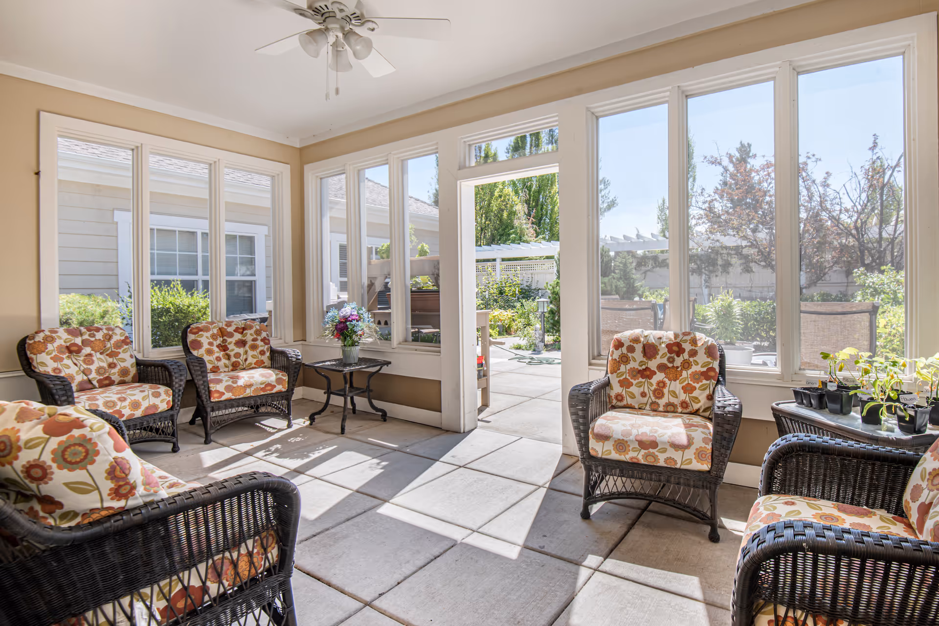 A sunlit indoor seating area with wicker chairs that have floral patterned cushions. The room has large windows and a glass door that opens to an outdoor garden patio with greenery and trees visible outside. There is a small table with a vase of flowers and another table with small potted plants.